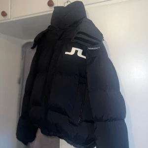 Pufferjacka från J.Lindeberg - Säljer en svart pufferjacka från J.Lindeberg med stor huva och Stort vitt logotryck på bröstet. Jackan har snörning nedtill, dragkedja, 2 inre & ute fickor och långa ärmar. Perfekt för dig som vill ha en varm och stilren look under vintern. Passar för båda könen och som L/M