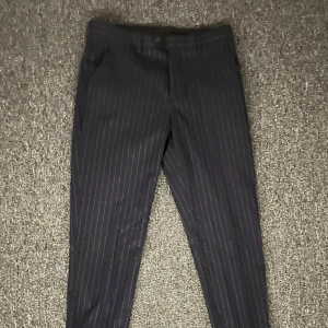 Mörkgrå randiga kostymbyxor devils advocate  - Snygga mörkgrå kostymbyxor med smala vita ränder. Byxorna har klassisk rak passform, bälteshällor och sidofickor fram samt knäppning med knapp och dragkedja. Perfekta för en stilren look och passar till skjorta eller t-shirt.