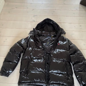 Svart pufferjacka från Polo Ralph Lauren - Säljer en svart, glansig pufferjacka från Polo Ralph Lauren med huva och dragkedja. Jackan har två sidofickor med dragkedja och den klassiska röda Polo-loggan broderad på bröstet. Riktigt fin jacka säljer då jag fick två st jackor i present så det blev tyvärr ett missförstånd. Kom med prisförslag om ni inte gillar priset. Alla taggar och allting sitter kvar då jag endast testat den