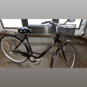 Yosemite City Bike - Stilren svart Yosemite damcykel med Shimano-växlar, cykelkorg och pakethållare. Cykeln är i gott skick med mindre yttre slitage, perfekt för stadskörning. Sadel och handtag ser hela ut, korgen har några löv men är intakt. Stabil och pålitlig cykel för vardagsbruk.
