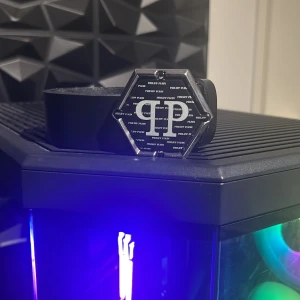 Svart bälte från Philipp Plein - Snyggt svart bälte från Philipp Plein med en unik sexkantig spänne i silverfärg. Spännet har logotyp och mönster med texten 'PHILIPP PLEIN' över hela ytan. Bältet har en stilren och modern look som passar till jeans eller kostym.