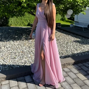 Glittrig rosa långklänning med slits - Säljer en rosa långklänning med glittrigt tyg och v-ringad omlottliv. Klänningen har ärmlös design och en snygg slits framtill. Använd endast några timmar, inga fel på den helt ny. Kan användas för bal, fest osv. 