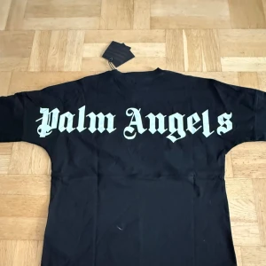 Svart långärmad tröja Palm Angels - Svart långärmad tröja från Palm Angels med stor vit logga längs ryggen och mindre logga tryckt vid halsen. Tröjan har rund halsringning och är gjord i mjuk bomull. Perfekt för dig som gillar streetwear och vill sticka ut med en ikonisk look.