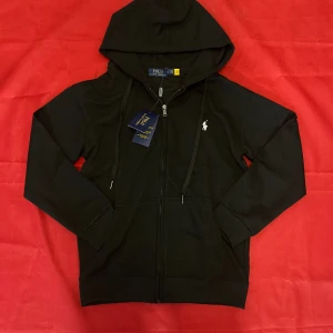 Svart hoodie från Polo Ralph Lauren - Svart hoodie från Polo Ralph Lauren med dragkedja framtill, huva med snörning och klassisk vit logga på bröstet. Mjuk bomullskvalitet och ribbade muddar. Perfekt för en chill och stilren look.