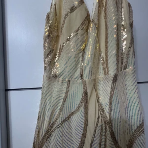 Champagnefärgad långklänning med paljetter - Långklänning i champagnefärg med glittriga paljettdetaljer i guld och mönster av böljande linjer. Djup v-ringning och elegant passform som ger en lyxig känsla. Perfekt för dig som vill sticka ut med en glamorös look.