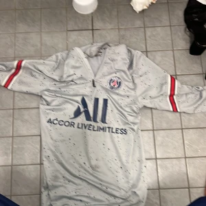 PSG träningströja grå Jordan - Snygg grå träningströja från PSG och Jordan med halv dragkedja, svarta prickdetaljer och röda samt vita ränder på ärmarna. PSG-logga på bröstet och stor 'All Accor Live Limitless'-logga framtill. Perfekt för fotbollsträning eller gym.