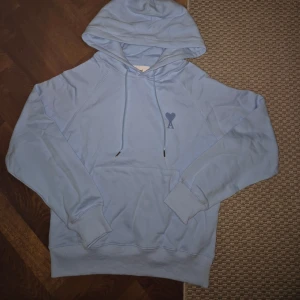  Ami hoodie - Säljer en snygg ljusblå hoodie från Ami Paris med klassisk huva och snörning. Tröjan har en broderad logga med hjärta och A på bröstet samt ribbade muddar. Perfekt för en chill och stilren look.