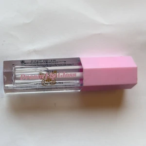 Dream Lip Gloss med rosa lock - Säljer ett Dream Lip Gloss med transparent glans och söt fjäril på förpackningen. Kommer i en rektangulär tub med ett ljusrosa lock. Perfekt för dig som vill ha glansiga och mjuka läppar med en touch av söt design. Endast testad på handen 