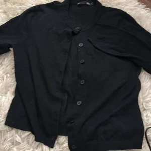 Svart kofta  - Svart kofta från 157 med ribbad krage och knappar framtill. Klassisk design med långärmade och mjukt material som känns skönt mot huden. Perfekt att slänga på sig när det är lite kyligt ute. Enkel och stilren, passar till det mesta.