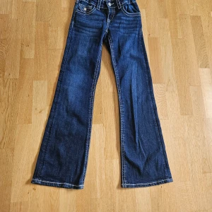 Blå bootcut jeans från Gina Tricot - Snygga mörkblå jeans från Gina Tricot i storlek 152. Modellen har bootcut-ben, klassiska fickor fram och bak med dekorativa knappar på bakfickorna. Jeansen har kontrastsömmar och är tillverkade i jeansmaterial med normal passform.