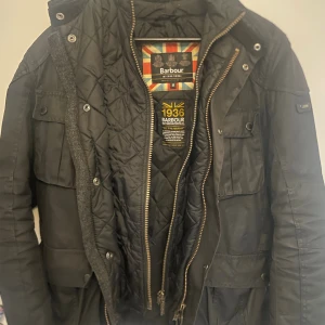 Svart Barbour International jacka M - Säljer en svart Barbour International jacka i vaxad bomull med quiltat foder. Jackan har flera stora fickor med knappar, hög krage och dragkedja framtill. Klassisk och stilren modell som funkar perfekt till höst och vinter. Storlek medium.