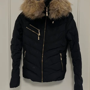 Svart pufferjacka Hollies fuskpäls XS - Svart pufferjacka från Hollies, stor huva med fluffig fuskpälssom går att ta av och leopardmönstrat innerfoder. Har en liten fläck på framsidan på magen som man ser på bild 1 men annars är den okej