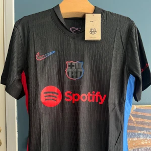 Svart FC Barcelona Nike fotbollströja - Säljer en svart FC Barcelona fotbollströja från Nike med röd Spotify-logga på bröstet och UNHCR-tryck på ryggen. Tröjan har korta ärmar, subtilt mönster i tyget och detaljer i blått och rött på sidorna. Materialet är lätt och andas, perfekt för match eller träning.