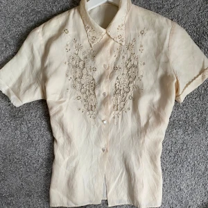 Beige blommig skjorta med spetsdetaljer - Supersöt beige skjorta med korta ärmar och spetsbroderier i blommigt mönster framtill. Skjortan har knappar och en krage med fina spetsdetaljer. Materialet känns lent och har en lätt glans, vilket ger en lyxig vibe. Perfekt för dig som gillar vintage och romantiska detaljer. Passar S/M.