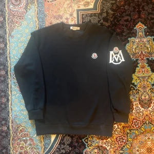 Svart sweatshirt från Moncler - Svart sweatshirt från Moncler med klassisk rund hals och ribbade muddar. Tröjan har Moncler-logga på bröstet och en stor vit M-detalj med logga på ena ärmen. Perfekt för dig som gillar stilrena streetwear-plagg.