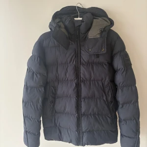 Svart pufferjacka från G-Star RAW - Säljer en svart pufferjacka från G-Star RAW med huva och dragkedja framtill. Jackan har quiltad design, två fickor med knappstängning och en snygg patch på ärmen. Perfekt för kalla dagar och riktigt skön att bära.