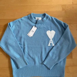 Ljusblå stickad tröja från Ami  - Snygg ljusblå stickad tröja från Ami Alexandre Mattiussi med vit hjärtdetalj och A på bröstet. Tröjan har rund hals, långa ärmar och breda ribbade muddar vid ärmslut och nederkant. Perfekt för dig som gillar stilrena och coola detaljer. Storlek S men passar Xs