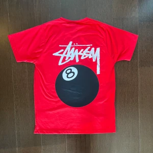 Röd Stüssy t-shirt med 8-ball print - Snygg röd t-shirt från Stüssy med stort 8-ball tryck och logga på ryggen samt mindre print på bröstet. Klassisk rund hals och korta ärmar. Materialet är mjuk bomull som känns skönt mot huden. Perfekt för dig som gillar streetwear och vill sticka ut.