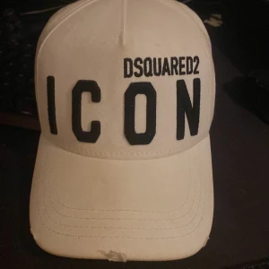 Vit Dsquared2 ICON keps - Snygg vit keps från Dsquared2 med stor svart 'ICON'-text framtill och broderad logga. Klassisk böjd skärm och justerbar passform. Perfekt för dig som vill sticka ut med en trendig accessoar.
