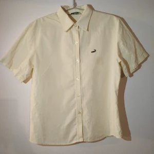 Gul kortärmad skjorta från Lacoste - Klassisk Gul skjorta från Lacoste med korta ärmar och diskret rutmönster. Skjortan har knappar framtill och den ikoniska krokodilloggan broderad på bröstet. Perfekt för en clean och stilren look. Liten olja fläck på högra sidan, enkel att ta bort
