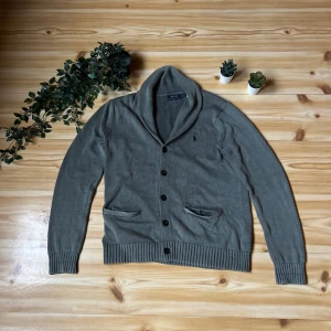 Grå kofta från Polo Ralph Lauren - Snygg grå kofta från Polo Ralph Lauren med klassisk sjalkrage och svarta knappar framtill. Stickad i mjuk bomull och har två fickor fram samt den ikoniska broderade loggan på bröstet. Perfekt för lager-på-lager och chill stil. pris kan diskuteras, tveka inte på att höra av er, mvh karl