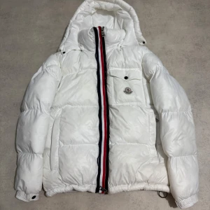 Vit Moncler pufferjacka med huva - Säljer en riktigt fet vit pufferjacka från Moncler med huva och coola Moncler-detaljer. Jackan har en röd, vit och blå rand längs dragkedjan och man kan dra upp kedjan utan som färgerna, bröstficka med knapp och broderad logga. Perfekt för kalla dagar när du vill sticka ut och hålla värmen.