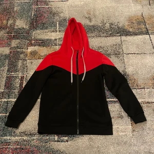 Svart och röd hoodie med dragkedja - Snygg hoodie i svart och rött med kontrastfärg över axlar och huva. Tröjan har långa ärmar, grå snören i huvan och hel dragkedja framtill. Perfekt för chill dagar eller när du vill ha en sportig look. Mjuk och bekväm med klassisk passform.