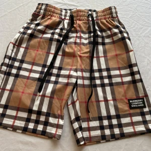 Rutig Burberry shorts med snörning - Snygga shorts Burberry med klassiskt rutigt mönster i beige, svart, vitt och röda detaljer. De har elastisk midja med svart snörning och en patch med Burberry London England på benet. Perfekta för sommaren och ger en lyxig vibe till din outfit.