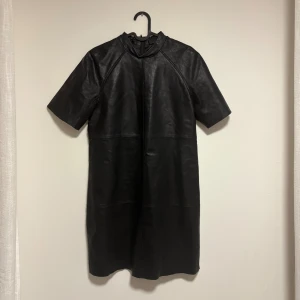 Svart skinnklänning med dragkedja - Säljer en svart kort skinnklänning med rak passform och korta ärmar. Klänningen har en hög krage och dragkedja baktill. Utsidan är i äkta läder och insidan är fodrad med polyester för extra komfort. Perfekt för dig som gillar en edgy och stilren look.