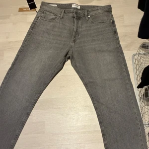 Grå jeans från Jack & Jones, straight fit - Säljer ett par gråa jeans från Jack & Jones i modellen JJICHRIS. Klassisk femficksdesign, rak passform och normal midja. Jeansen är tillverkade i denim och har snygga detaljer som metallnitar och läderpatch bak. Perfekt för dig som gillar stilrena och tidlösa jeans.