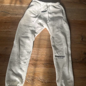 Vita mjukisbyxor Essentials Fear of God - Snygga vita mjukisbyxor från Essentials Fear of God med tryck på benet och elastisk midja med snörning. Byxorna har en relaxed passform och är gjorda i mjukt bomullsmaterial, perfekta för chill dagar. Stilsäkert streetwear-plagg med clean look.