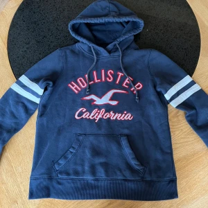 Marinblå Hollister hoodie med tryck - Snygg marinblå hoodie från Hollister med rött och vitt tryck på bröstet och vita ränder på ärmarna. Tröjan har huva med snörning, magficka och ribbade muddar. Perfekt för chill dagar och har en sportig vibe.