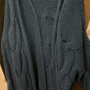 Mörkblå stickad kofta - Säljer en chunky mörkblå stickad kofta med grova kabelstickade detaljer. Koftan har en öppen front och är perfekt för att slänga över en t-shirt när det är kyligt. Den har lång ärm och en mysig, oversized känsla.