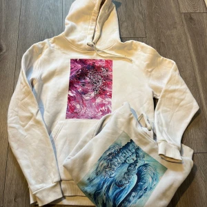 Vit hoodie med konstmotiv  - Två vita hoodies från The Cool Elephant i storlek L, med unika konstmotiv på bröstet – en med rosa och rött ansikte med blommor, den andra med blått och turkost motiv av en kvinna. Båda har huva, snörning och magficka. Perfekt för dig som gillar coola prints och streetstyle.