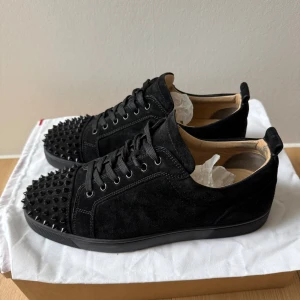 Christian Louboutin junior spike  - Svarta sneakers i mocka från Christian Louboutin med ikoniska röda sulor och coola svarta nitar på tån. Skorna har snörning och en platt sula, vilket ger en edgy vibe. Perfekta för dig som vill sticka ut med lyxig streetstyle. Str. 43