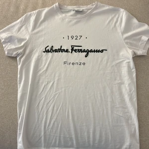 Vit t-shirt Salvatore Ferragamo M - Vit t-shirt från Salvatore Ferragamo med svart tryck framtill där det står '1927 Salvatore Ferragamo Firenze'. Klassisk rund hals och korta ärmar. Tillverkad i 100% mjuk bomull, perfekt för en clean och stilren look.