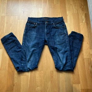 Blå jeans från Nudie Jeans, W30 L32 - Säljer ett par klassiska blå jeans från Nudie Jeans med snygg tvätt och kontrastsömmar. Modellen har fem fickor, raka ben och normal passform. Materialet är slitstark bomullsdenim och bakfickorna har Nudie Jeans ikoniska broderi. Byxorna är använda sparsamt då de inte passade mig. Modellen tape Ted. Pass formen är lik Lean Dean. Pris är diskuterbart 