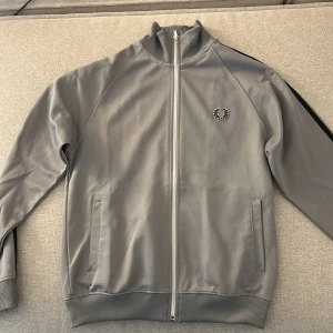 Grå track jacket från Fred Perry S - Snygg grå track jacket från Fred Perry med svart rand längs ärmarna och klassisk laurel-logga på bröstet. Jackan har hög krage, hel dragkedja och ribbade muddar. Perfekt för en sportig och stilren look.