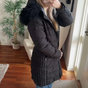 Svart lång dunjacka Rockandblue - Säljer en svart lång dunjacka från Rockandblue med quiltad design och stor huva med fluffig fuskpäls. Jackan har dragkedja, knappar och två fickor med dragkedja. Perfekt för kalla vinterdagar och riktigt snygg passform.