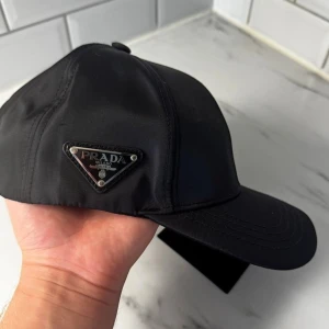 Svart keps från Prada i nylon - Svart keps från Prada med klassisk böjd skärm och justerbar rem baktill. Kepsen är tillverkad i glansigt nylonmaterial och har Pradas ikoniska metallogo i triangelform på sidan. Snygg och stilren accessoar som lyfter vilken outfit som helst.