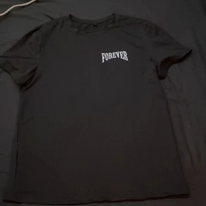 Svart t-shirt med Forever-tryck och ängel - Svart t-shirt med coolt Forever-tryck på bröstet och stor grafisk print på ryggen med en ängel, blå blixtar och textdetaljer. Klassisk passform och rund halsringning. Perfekt för dig som gillar streetstyle vibes. Passar även S 