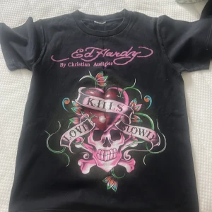 Svart Ed Hardy t-shirt med tryck - Svart t-shirt från Ed Hardy med stort färgglatt tryck framtill av dödskalle, hjärta och texten 'Love Kills Slowly'. Rosa logga både fram och bak. Klassisk passform och korta ärmar. Perfekt statement-plagg för dig som gillar edgy streetstyle.