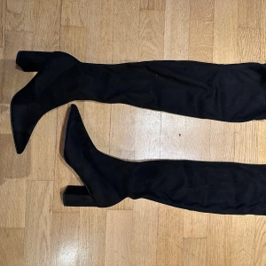 Svarta stövlar med klack från NA-KD - Säljer ett par svarta, höga stövlar från NA-KD med spetsig tå och bred klack. Skorna är i syntetmaterial med mockaliknande känsla och har en stilren, slimmad passform som går över knäet. Perfekta för att lyfta vilken outfit som helst.