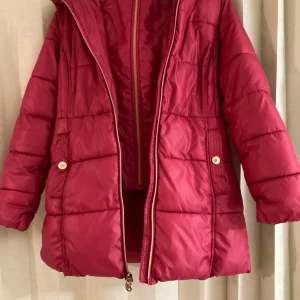 Röd pufferjacka från Michael Kors - Snygg röd pufferjacka från Michael Kors med guldig dragkedja och MK-logga. Jackan har huva, långa ärmar och quiltad design. Insidan är fodrad med mjuk fleece och har en dekorativ guldfärgad rand. Perfekt för kalla dagar och riktigt skön att bära.