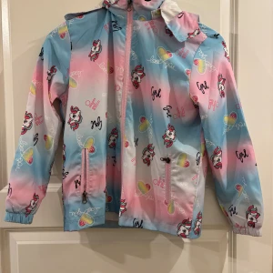 Pastellfärgad vindjacka med enhörningar  - Färgglad vindjacka från Shein i storlek 10Y med pastellrosa, blå och vita nyanser. Jackan har huva, dragkedja framtill och två fickor. Tryckt med enhörningar, hjärtan och text. Perfekt för dig som gillar lekfull stil och vill sticka ut. Pris kan diskuteras 