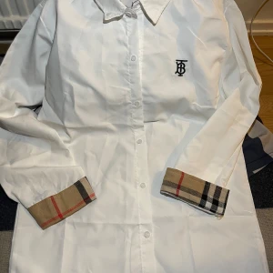 Vit Burberry skjorta - Vit skjorta från Burberry med klassisk krage, knäppning framtill och svart broderad logga på bröstet. Ärmsluten har den ikoniska beigea Burberry-rutan med röda och svarta inslag. Skjortan är långärmad och i bomull, perfekt för en clean och stilren look.