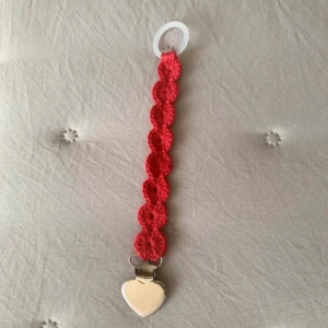 Rött virkat nappband med hjärtdetalj - Handgjort nappband i rött virkat material med rundade former och ett hjärtformat spänne i silverfärg. Bandet har en praktisk plastögla för att fästa nappen. Perfekt accessoar för att hålla koll på nappen med stil.
