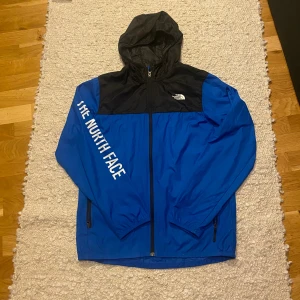 Blå och svart vindjacka The North Face - Snygg blå och svart vindjacka från The North Face med huva och hel dragkedja. Jackan har stora vita logotexter på bröstet och ärmen, samt två dragkedjefickor. Perfekt för dig som gillar streetwear och outdoor-vibe. Passar S size.