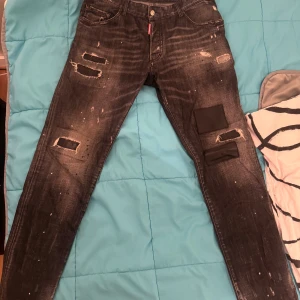 ÄKTA Svarta Dsquared2 Jeans - Säljer ett par svarta jeans från Dsquared2 med coola slitningar och patchdetaljer på benen. Jeansen har klassisk passform.  Storlek 54, liten i storlek, kort modell på byxorna. För mer bilder skriv privat 
