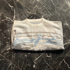 Vit och ljusblå stickad tröja - Vit stickad tröja med abstrakt mönster i ljusblått och grått. Tröjan har rund halsringning, ribbade muddar och är långärmad. Perfekt för dig som gillar en chill och stilren vibe med en twist av färg.